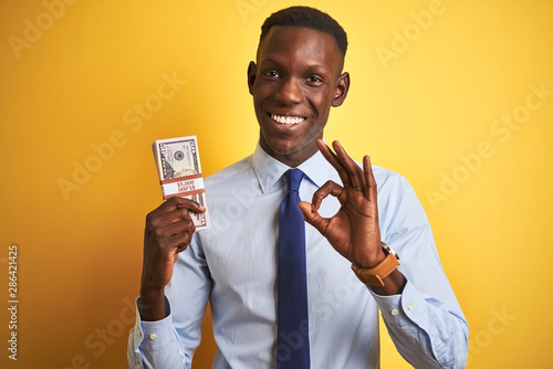 American businessman holding bunch of dollars standing over isolated yellow background doing ok sign with fingers, excellent symbol