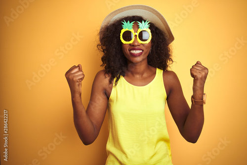 African american woman wearing funny pineapple sunglasses over isolated yellow background celebrating surprised and amazed for success with arms raised and open eyes. Winner concept.
