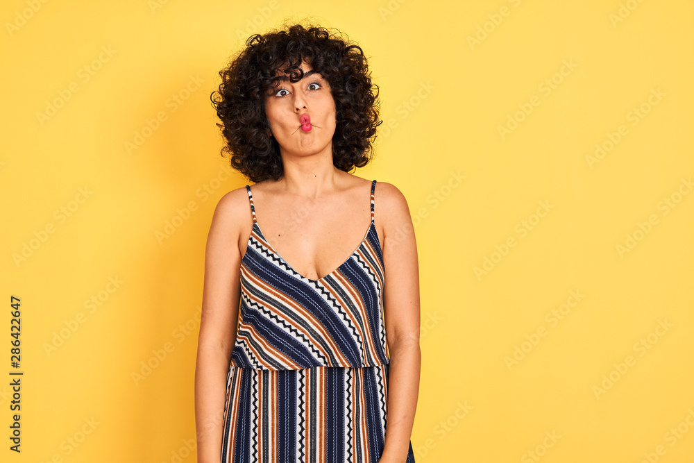 Arab woman with curly hair wearing striped colorful dress over isolated yellow background making fish face with lips, crazy and comical gesture. Funny expression. Arab woman with curly hair wearing striped colorful dress over isolated yellow background making fish face with lips, crazy and comical gesture. Funny expression.