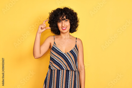 Arab woman with curly hair wearing striped colorful dress over isolated yellow background smiling and confident gesturing with hand doing small size sign with fingers looking and the camera.