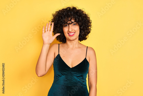 Young arab woman with curly hair wearing elegant dress over isolated yellow background showing and pointing up with fingers number five while smiling confident and happy.