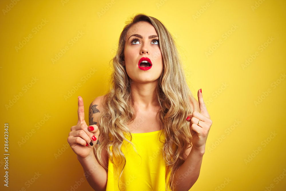 Young attactive woman wearing t-shirt standing over yellow isolated background amazed and surprised looking up and pointing with fingers and raised arms. Young attactive woman wearing t-shirt standing over yellow isolated background amazed and surprised looking up and pointing with fingers and raised arms.