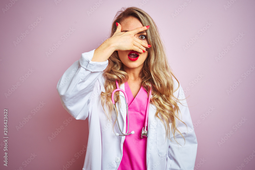 Young beautiful doctor woman using stethoscope over pink isolated background peeking in shock covering face and eyes with hand, looking through fingers with embarrassed expression. Young beautiful doctor woman using stethoscope over pink isolated background peeking in shock covering face and eyes with hand, looking through fingers with embarrassed expression.