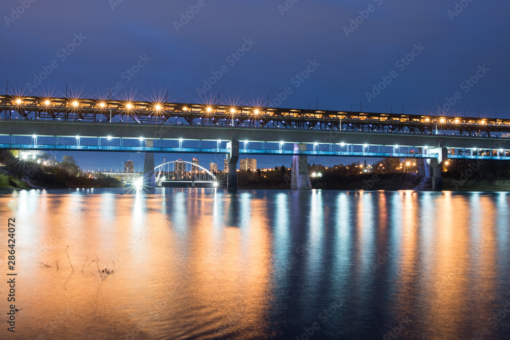 THE HIGH LEVEL BRIDGE AT 100, en la ciudad de Edmonton en Alberta Canadá, con un amanecer azul y luces rojas y naranjas que se reflejan en el río, mientras pasa el tren sobre el puente y tranvia.