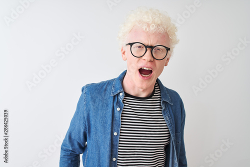 Young albino blond man wearing denim shirt and glasses over isolated white background angry and mad screaming frustrated and furious, shouting with anger. Rage and aggressive concept.