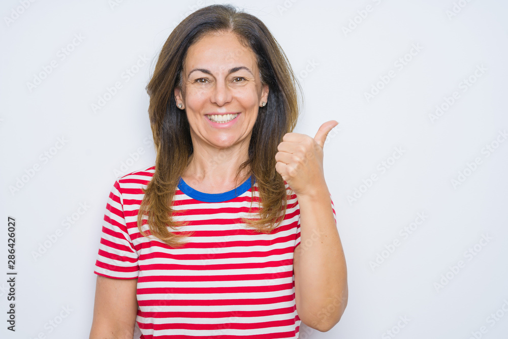 Middle age senior woman standing over white isolated background smiling with happy face looking and pointing to the side with thumb up. Middle age senior woman standing over white isolated background smiling with happy face looking and pointing to the side with thumb up.