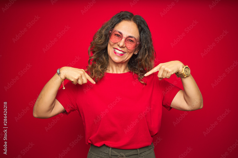 Middle age senior woman wearing cute heart shaped glasses over red isolated background looking confident with smile on face, pointing oneself with fingers proud and happy. Middle age senior woman wearing cute heart shaped glasses over red isolated background looking confident with smile on face, pointing oneself with fingers proud and happy.