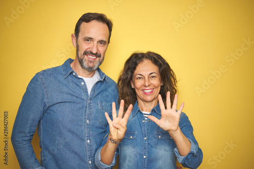Beautiful middle age couple together standing over isolated yellow background showing and pointing up with fingers number nine while smiling confident and happy.