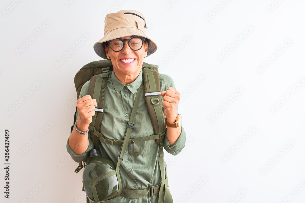 Middle age hiker woman wearing backpack canteen hat glasses over isolated white background excited for success with arms raised and eyes closed celebrating victory smiling. Winner concept. Middle age hiker woman wearing backpack canteen hat glasses over isolated white background excited for success with arms raised and eyes closed celebrating victory smiling. Winner concept.