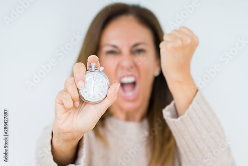 Middle age woman holding stopwatch isolated background annoyed and frustrated shouting with anger  crazy and yelling with raised hand  anger concept