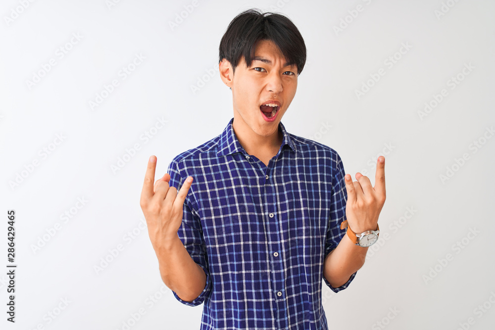 Young chinese man wearing casual blue shirt standing over isolated white background shouting with crazy expression doing rock symbol with hands up. Music star. Heavy concept. Young chinese man wearing casual blue shirt standing over isolated white background shouting with crazy expression doing rock symbol with hands up. Music star. Heavy concept.