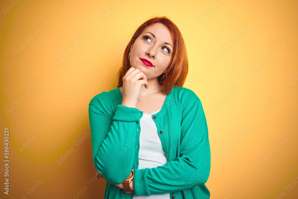 Youg beautiful redhead woman wearing winter green sweater over isolated yellow background with hand on chin thinking about question, pensive expression. Smiling with thoughtful face. Doubt concept. Youg beautiful redhead woman wearing winter green sweater over isolated yellow background with hand on chin thinking about question, pensive expression. Smiling with thoughtful face. Doubt concept.