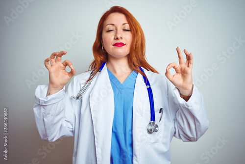 Young redhead doctor woman using stethoscope over white isolated background relaxed and smiling with eyes closed doing meditation gesture with fingers. Yoga concept.