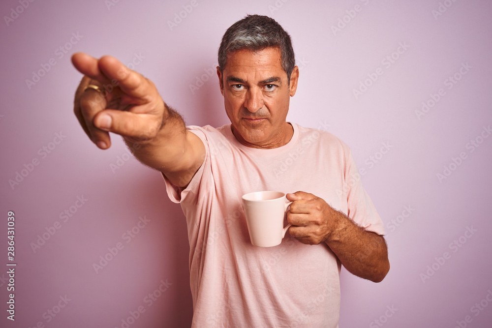 Handsome middle age man holding cup of coffee standing over isolated pink background pointing with finger to the camera and to you, hand sign, positive and confident gesture from the front Handsome middle age man holding cup of coffee standing over isolated pink background pointing with finger to the camera and to you, hand sign, positive and confident gesture from the front