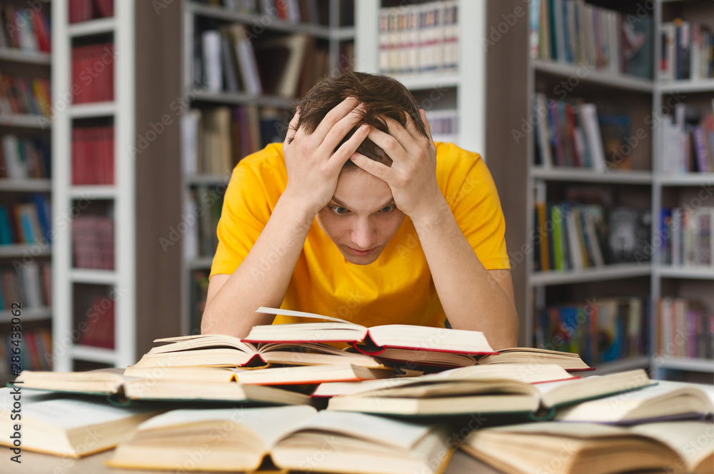 Exhausted guy holding his head and reading book in library Exhausted guy holding his head and reading book in library