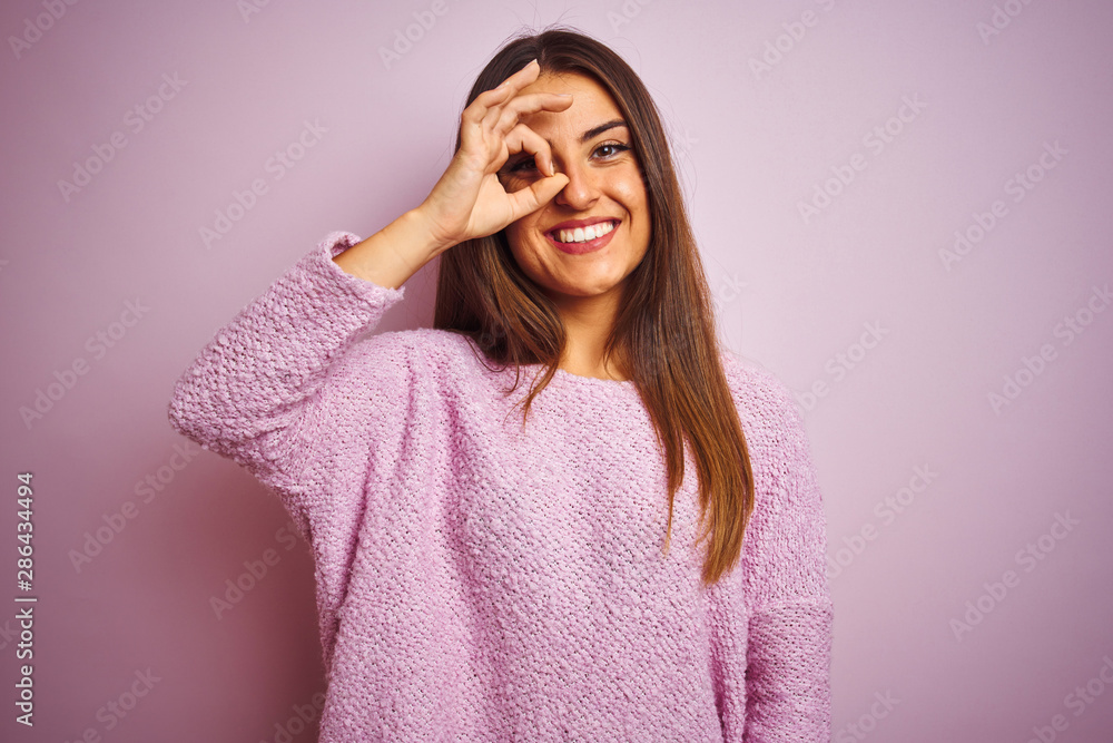 Young beautiful woman wearing casual sweater standing over isolated pink background doing ok gesture with hand smiling, eye looking through fingers with happy face. Young beautiful woman wearing casual sweater standing over isolated pink background doing ok gesture with hand smiling, eye looking through fingers with happy face.