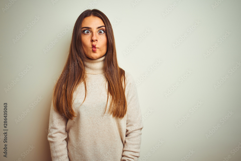 Obraz premium Young beautiful woman wearing winter turtleneck sweater over isolated white background making fish face with lips, crazy and comical gesture. Funny expression.