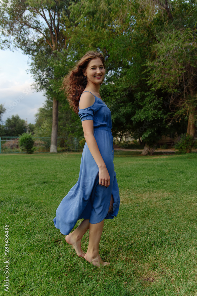 A young woman in a blue dress walks barefoot on the grass in the park A young woman in a blue dress walks barefoot on the grass in the park