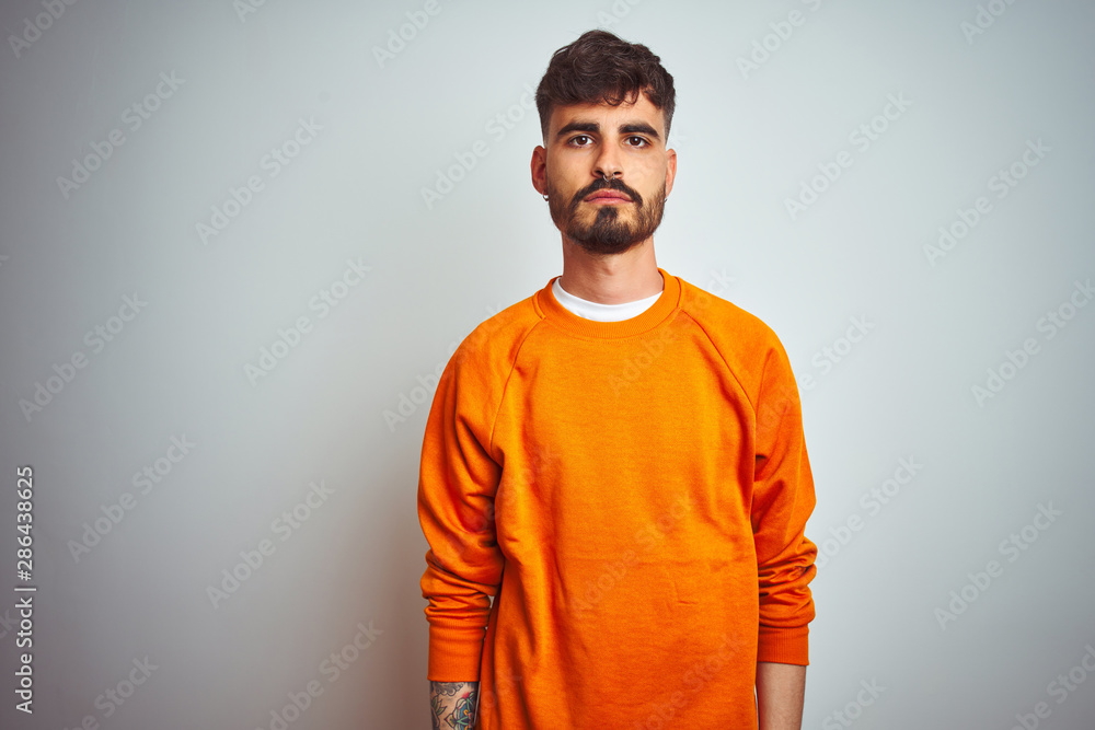 Young man with tattoo wearing orange sweater standing over isolated white background Relaxed with serious expression on face. Simple and natural looking at the camera. Young man with tattoo wearing orange sweater standing over isolated white background Relaxed with serious expression on face. Simple and natural looking at the camera.