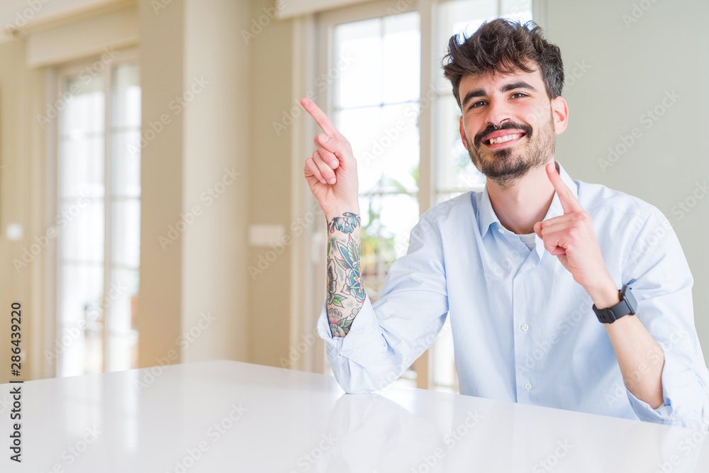 Young businesss man sitting on white table smiling and looking at the camera pointing with two hands and fingers to the side. Young businesss man sitting on white table smiling and looking at the camera pointing with two hands and fingers to the side.