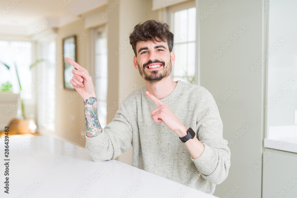 Young man wearing casual sweater sitting on white table smiling and looking at the camera pointing with two hands and fingers to the side.