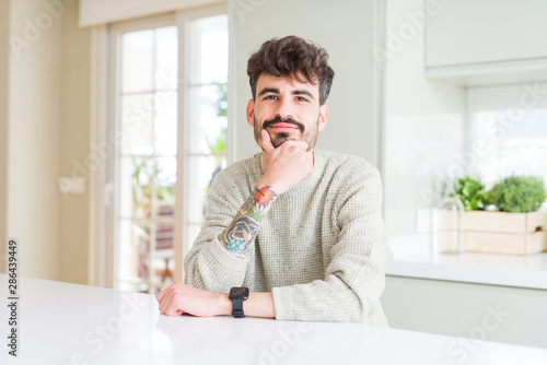 Young man wearing casual sweater sitting on white table looking confident at the camera with smile with crossed arms and hand raised on chin. Thinking positive.