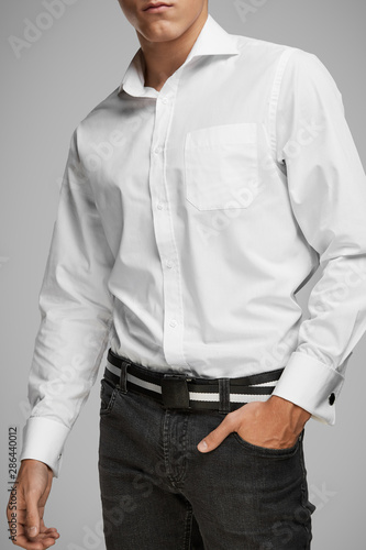 Cropped medium full shot of a young man dressed in a white buttoned shirt with a pocket and gray jeans with a belt. The black belt with a white stripe is made of textured fabric and equipped 