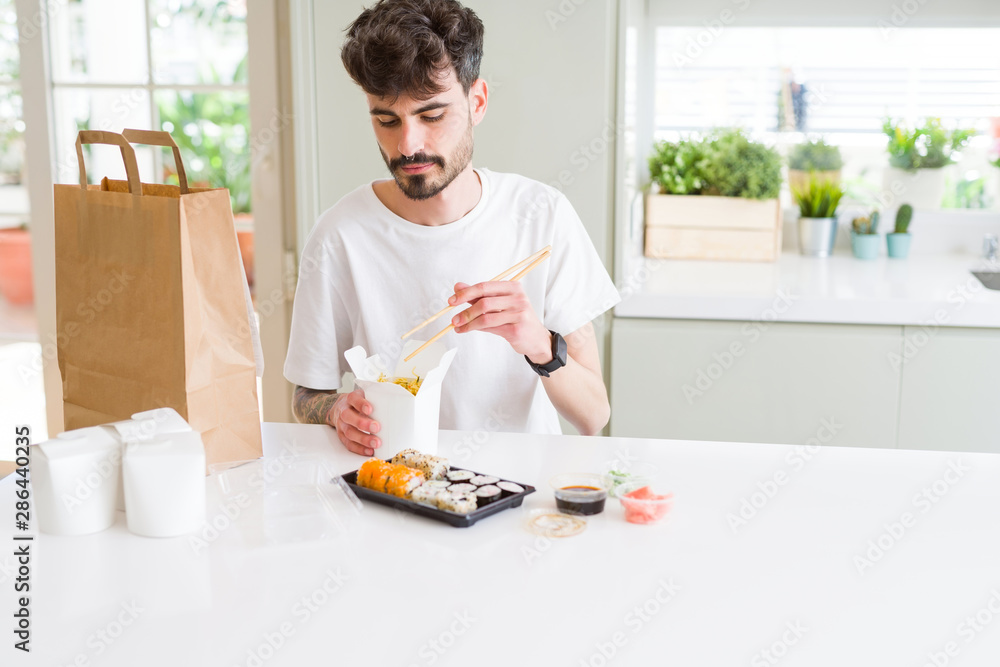 Young man eating sushi asian food and noodles using choopsticks from take away delivery Young man eating sushi asian food and noodles using choopsticks from take away delivery