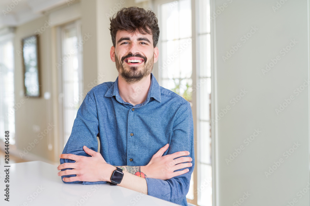 Young man wearing casual shirt sitting on white table happy face smiling with crossed arms looking at the camera. Positive person. Young man wearing casual shirt sitting on white table happy face smiling with crossed arms looking at the camera. Positive person.
