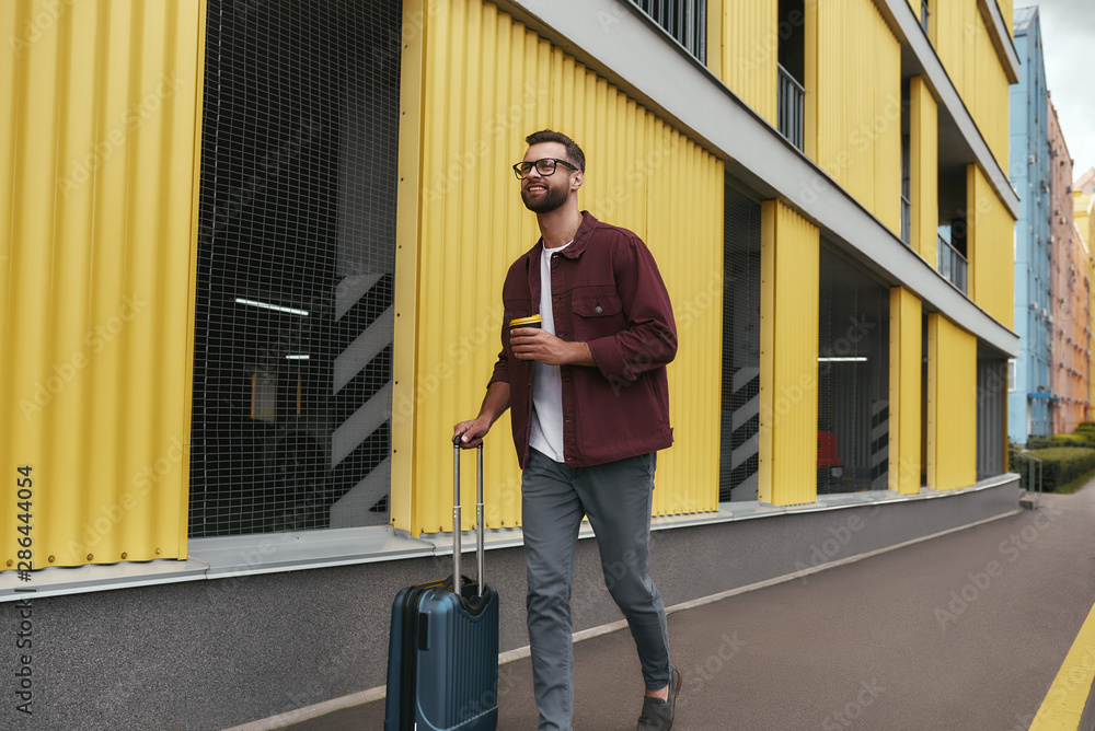 Travel mood. Handsome smiling bearded man in casual wear and eyeglasses holding a disposable cup and carrying his luggage while walking through the city street