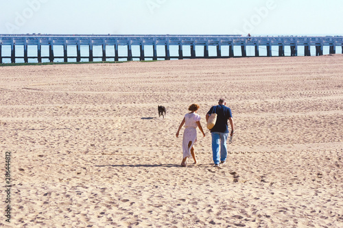 A married couple, a woman in a white dress, and a man walk barefoot along the beach in Ostend. Belgium. Early in the morning by the sea. Wooden bridge against the sky. Traveling together.