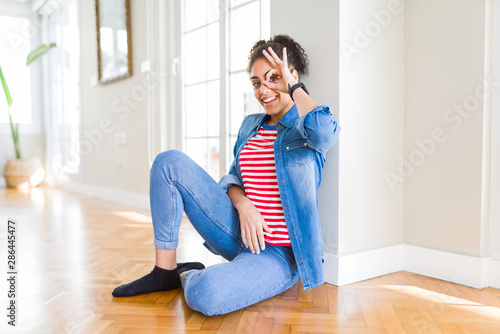 Beautiful young african american woman with afro hair sitting on the floor doing ok gesture with hand smiling, eye looking through fingers with happy face.