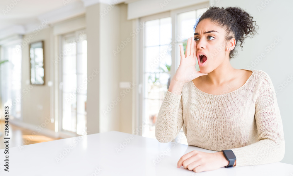 Beautiful young african american woman with afro hair sitting on table at home shouting and screaming loud to side with hand on mouth. Communication concept. Beautiful young african american woman with afro hair sitting on table at home shouting and screaming loud to side with hand on mouth. Communication concept.