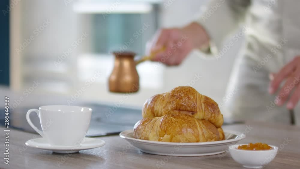 Close-up shot of unrecognizable man picking up cezve from electric stove, pouring steaming fresh coffee into white cup on saucer, and croissants and jam on table nearby Close-up shot of unrecognizable man picking up cezve from electric stove, pouring steaming fresh coffee into white cup on saucer, and croissants and jam on table nearby