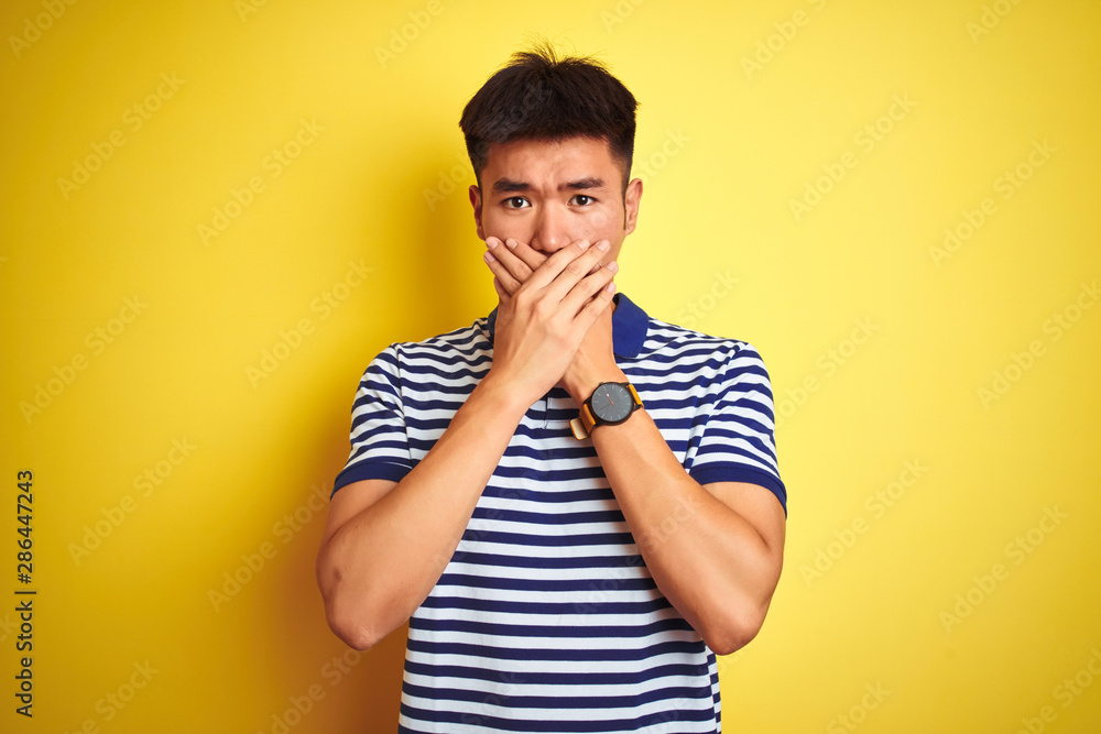Young asian chinese man wearing striped polo standing over isolated yellow background shocked covering mouth with hands for mistake. Secret concept. Young asian chinese man wearing striped polo standing over isolated yellow background shocked covering mouth with hands for mistake. Secret concept.