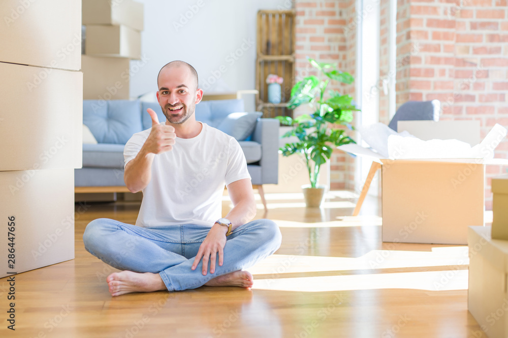 Young bald man sitting on the floor around cardboard boxes moving to a new home doing happy thumbs up gesture with hand. Approving expression looking at the camera with showing success. Young bald man sitting on the floor around cardboard boxes moving to a new home doing happy thumbs up gesture with hand. Approving expression looking at the camera with showing success.