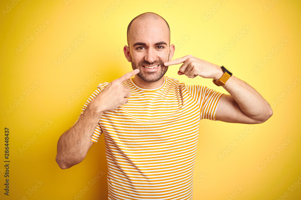 Young bald man with beard wearing casual striped t-shirt over yellow isolated background smiling cheerful showing and pointing with fingers teeth and mouth. Dental health concept. Young bald man with beard wearing casual striped t-shirt over yellow isolated background smiling cheerful showing and pointing with fingers teeth and mouth. Dental health concept.