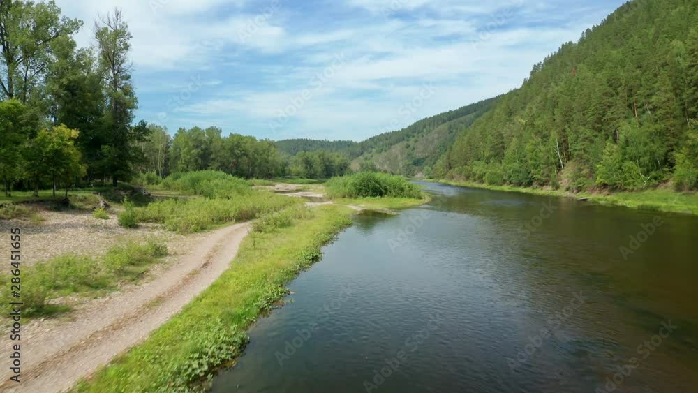 A copter flies low over the river toward the shore. There is a country road near the shore. The opposite shore is steep with dense vegetation. A copter flies low over the river toward the shore. There is a country road near the shore. The opposite shore is steep with dense vegetation.