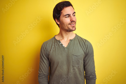 Young handsome man wearing casual t-shirt standing over isolated yellow background looking away to side with smile on face, natural expression. Laughing confident.