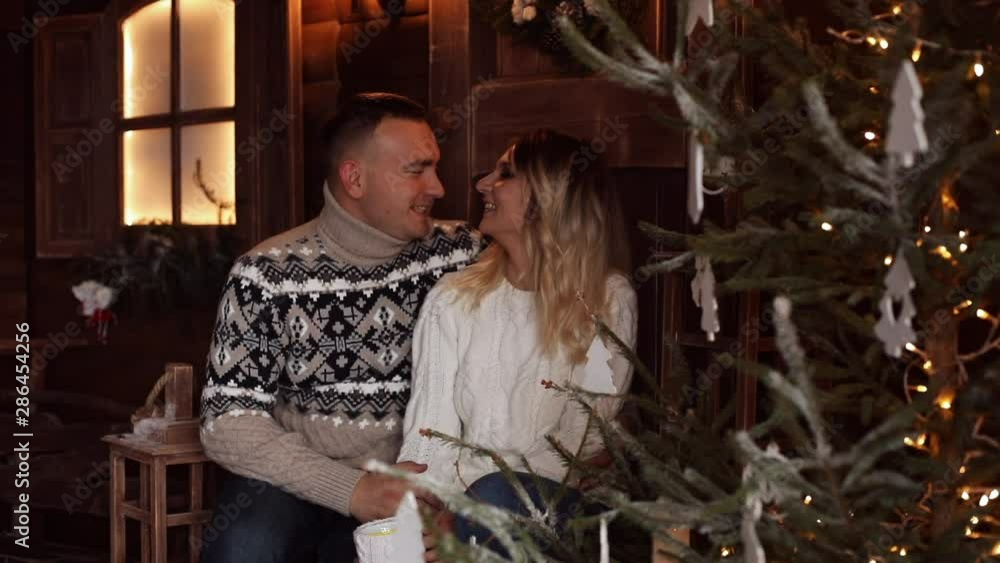 Happy and loving couple in sweaters and hats kissing while sitting on bench under festive golden confetti. They are drinking hot beverage and sitting in beautifully decorated room with Christmas tree. Happy and loving couple in sweaters and hats kissing while sitting on bench under festive golden confetti. They are drinking hot beverage and sitting in beautifully decorated room with Christmas tree.