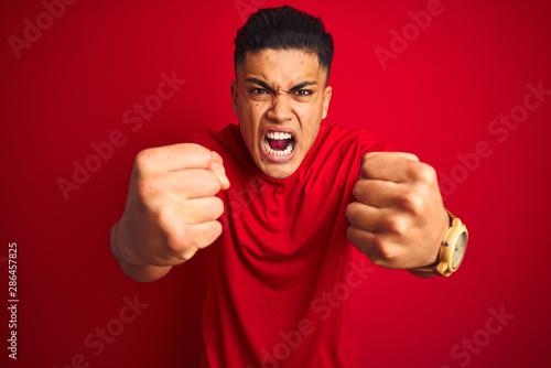 Bild auf Leinwand Young brazilian man wearing t-shirt standing over isolated red background angry and mad raising fists frustrated and furious while shouting with anger
