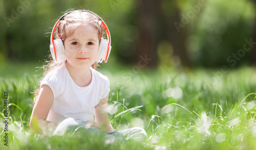 Cute little girl is listening to music in the park. Family outdoor lifestyle. Happy small kid in headphones sitting on green grass. Beauty nature at summer. Childhood happiness.  Children day.