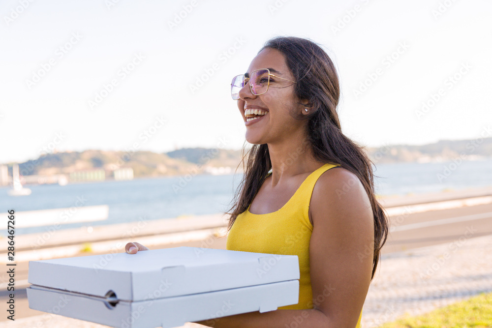 Happy Latin woman walking to meet friends and carrying pizza. Beautiful young woman walking outdoors, holding pizza boxes, looking into vacancy, smiling and laughing. Food delivery concept
