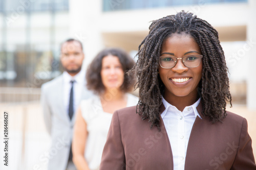 Cheerful young African American businesswoman. Portrait of confident female boss smiling at camera while standing with multiethnic colleagues, selective focus. Leadership concept