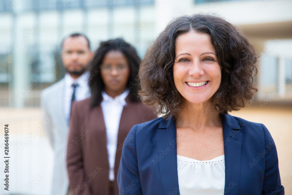Middle aged businesswoman smiling at camera. Portrait of cheerful professional businesswoman standing with colleagues, selective focus. Leadership concept Middle aged businesswoman smiling at camera. Portrait of cheerful professional businesswoman standing with colleagues, selective focus. Leadership concept