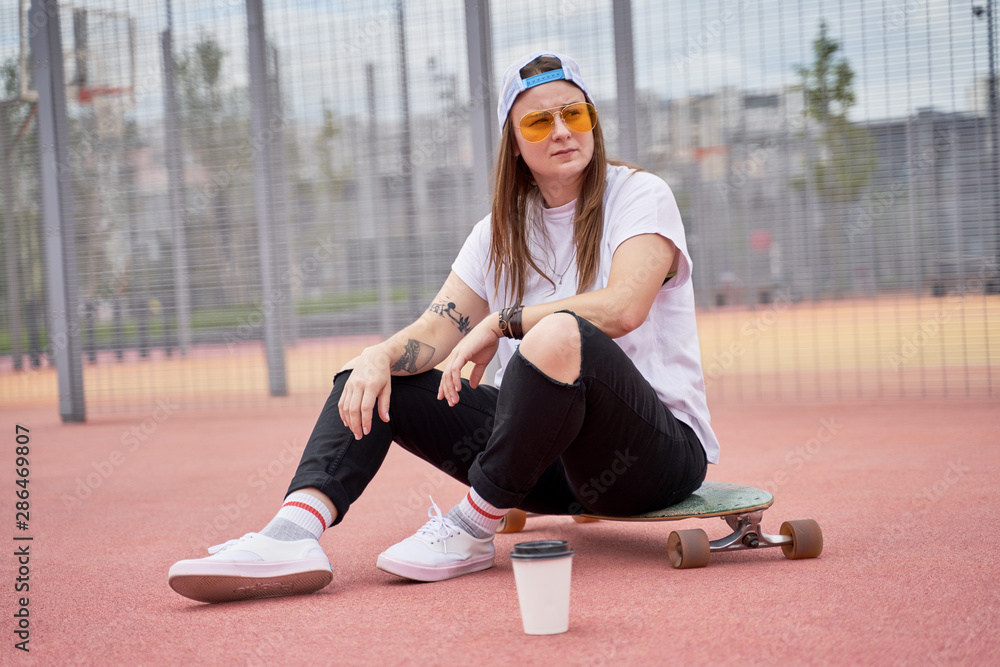 Fototapeta premium Image of athletic brunette girl in sunglasses sitting on skateboard on playground on summer day