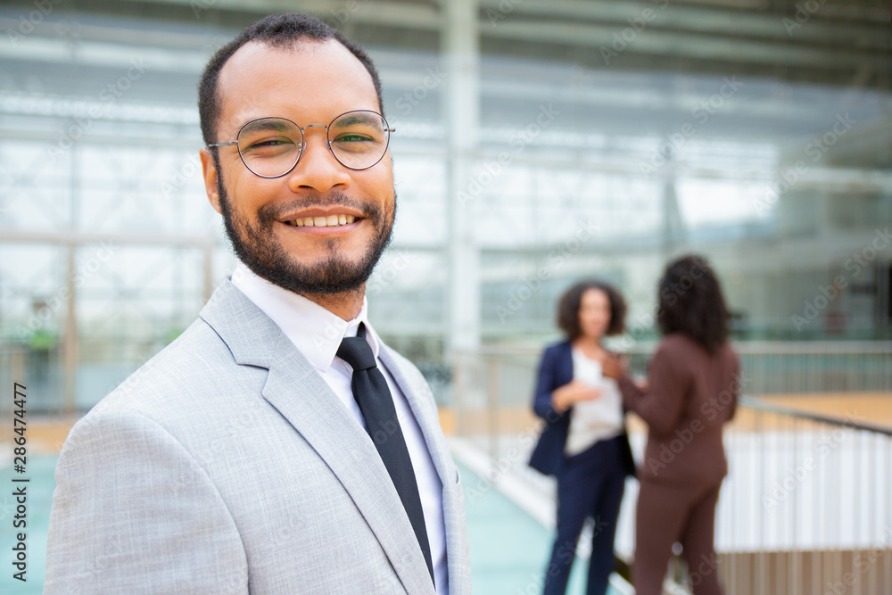 Cheerful handsome businessman. Portrait of professional young African American businessman in eyeglasses smiling at camera while female colleagues standing behind. Leader concept Cheerful handsome businessman. Portrait of professional young African American businessman in eyeglasses smiling at camera while female colleagues standing behind. Leader concept