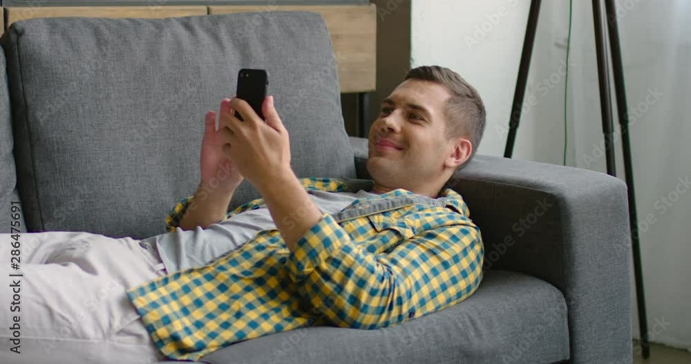 Handsome man is lying on sofa, using his gadget, smartphone, browsing, looking at screen, having nice time at home. Caucasian. Slow motion, 4K. Shot on RED camera. Handsome man is lying on sofa, using his gadget, smartphone, browsing, looking at screen, having nice time at home. Caucasian. Slow motion, 4K. Shot on RED camera.