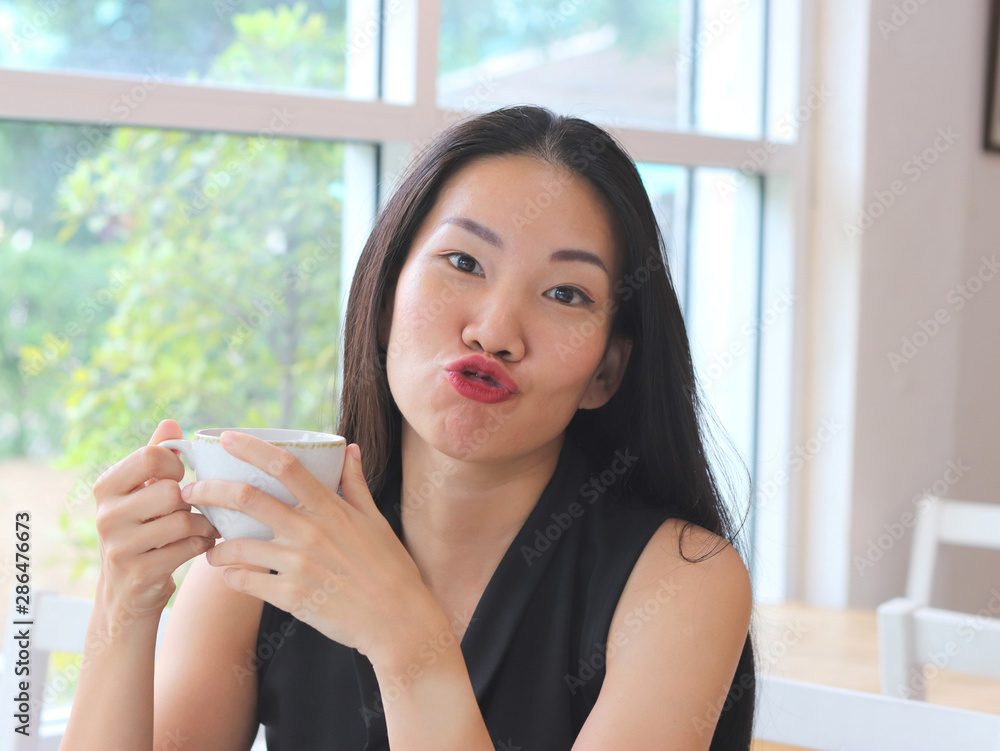 Asian woman making kissing and funny face with cup of coffee in her hands.  Stock Photo | Adobe Stock