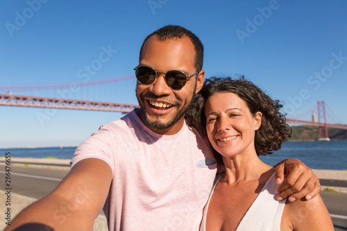 Couple of joyful tourists taking selfie on city promenade. Happy man and woman standing outside, holding smartphone and hugging. Happy couple concept